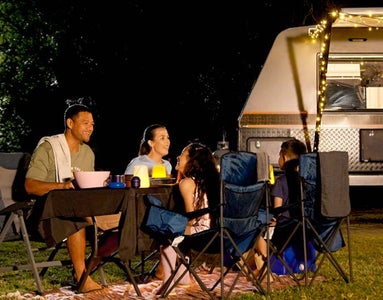 Family gathered around a camp table enjoying a meal at nighttime, in front of a caravan